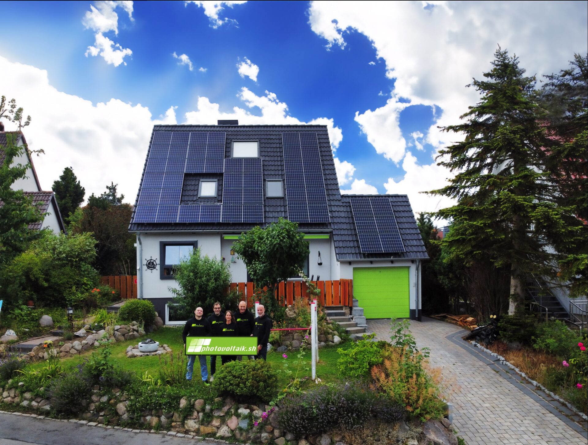 Vier Menschen mit einem grünen Schild mit der Aufschrift photovoltaik stehen vor einem weißen Haus mit Solarzellen auf dem Dach, einem grünen Garagentor und einem gepflegten Garten. Der Himmel ist teilweise bewölkt.